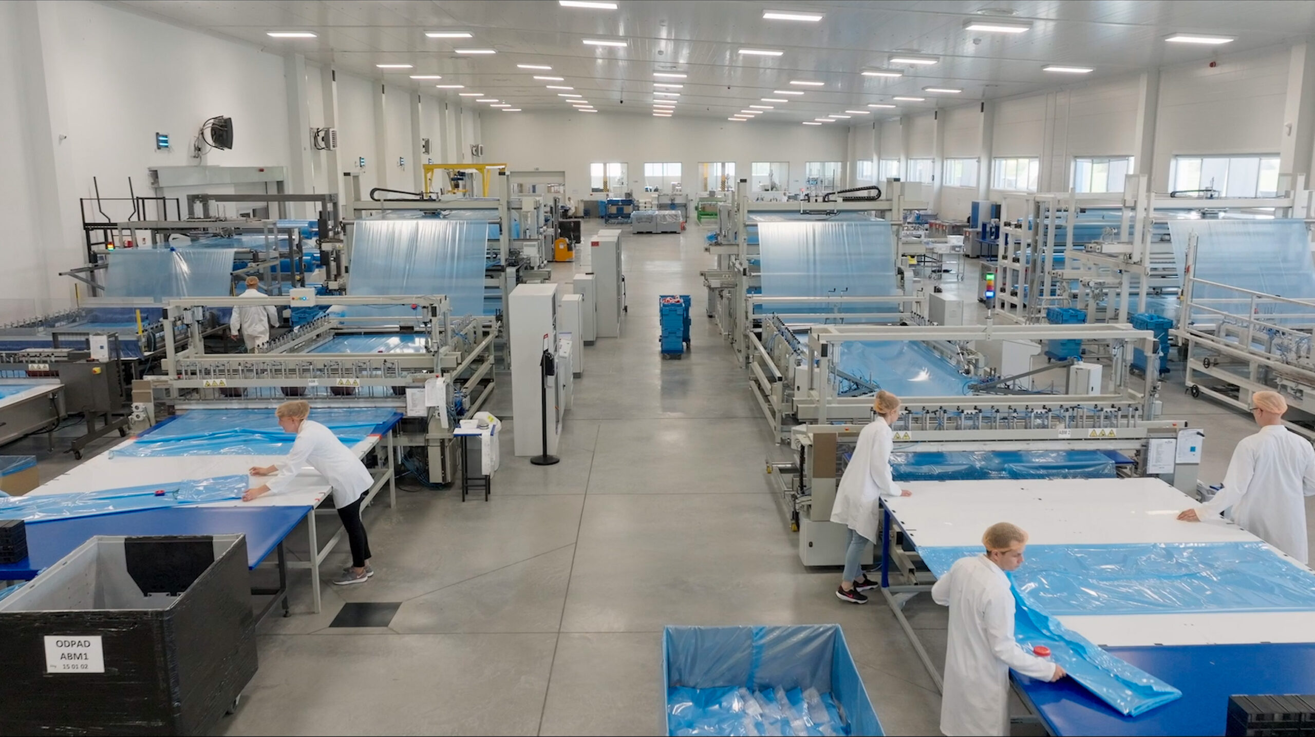 Nowoczesna hala produkcyjna firmy Peak Packaging z liniami do wytwarzania folii i opakowań higienicznych. Pracownicy w białych fartuchach nadzorują proces pakowania i kontroli jakości.